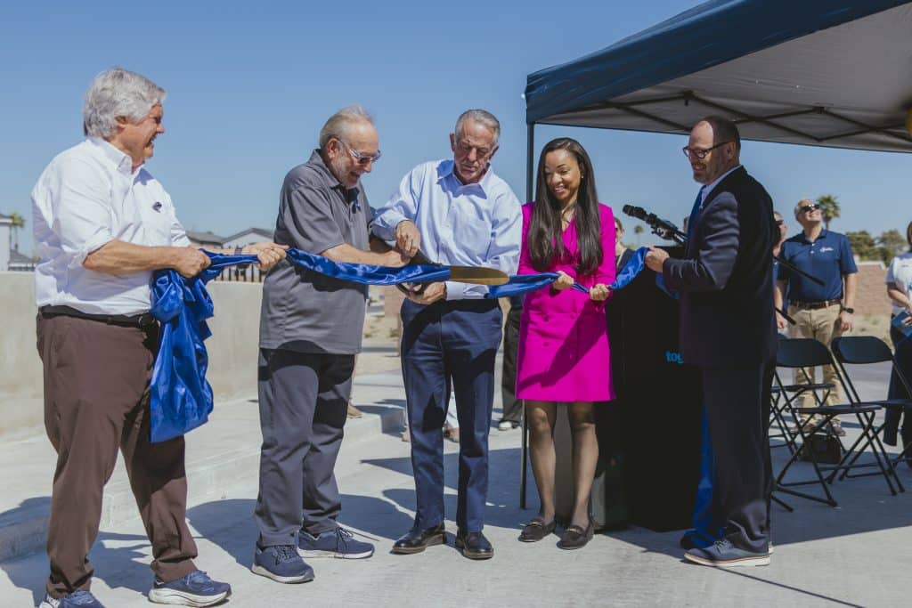Ribbon-cutting ceremony at Paradise Trails in Las Vegas, where local and state leaders use oversized scissors to cut a blue ribbon in celebration of the Richard H. Plaster Bridge opening and AB 540 housing funding; officials stand outdoors near a podium and canopy under a clear blue sky.