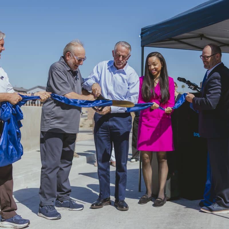 Ribbon-cutting ceremony at Paradise Trails in Las Vegas, where local and state leaders use oversized scissors to cut a blue ribbon in celebration of the Richard H. Plaster Bridge opening and AB 540 housing funding; officials stand outdoors near a podium and canopy under a clear blue sky.